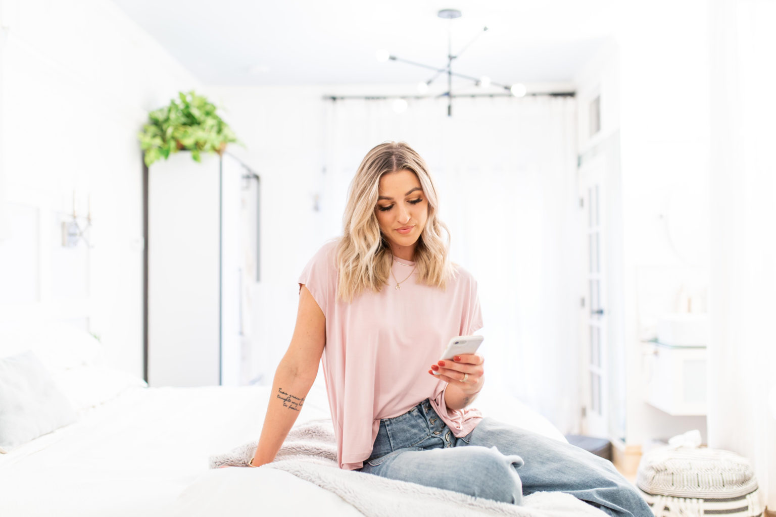 A woman is sitting on a bright white bed in a modern room looks down at her phone while checking something on the screen.
