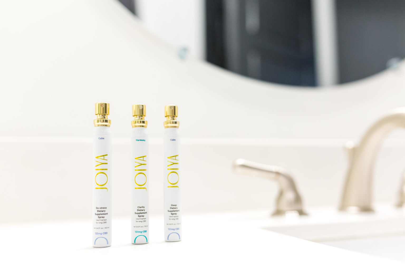 Three white and gold JOIYA dietary supplement spray bottles labeled Calm, Harmony, and Sleep stand on a bathroom counter near a sink.