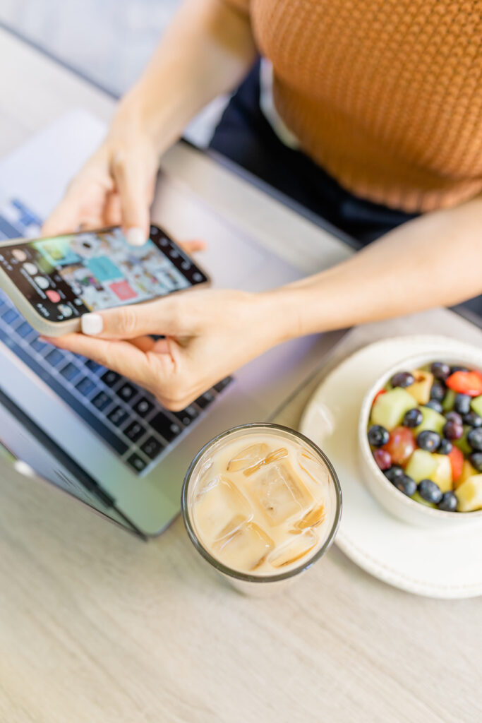 holding cell phone over laptop with iced coffee and bowl of fruit