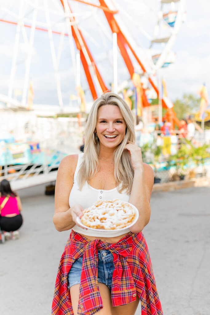 woman smiling posing for a summer lifestyle photoshoot at the fair holding a funnel cake