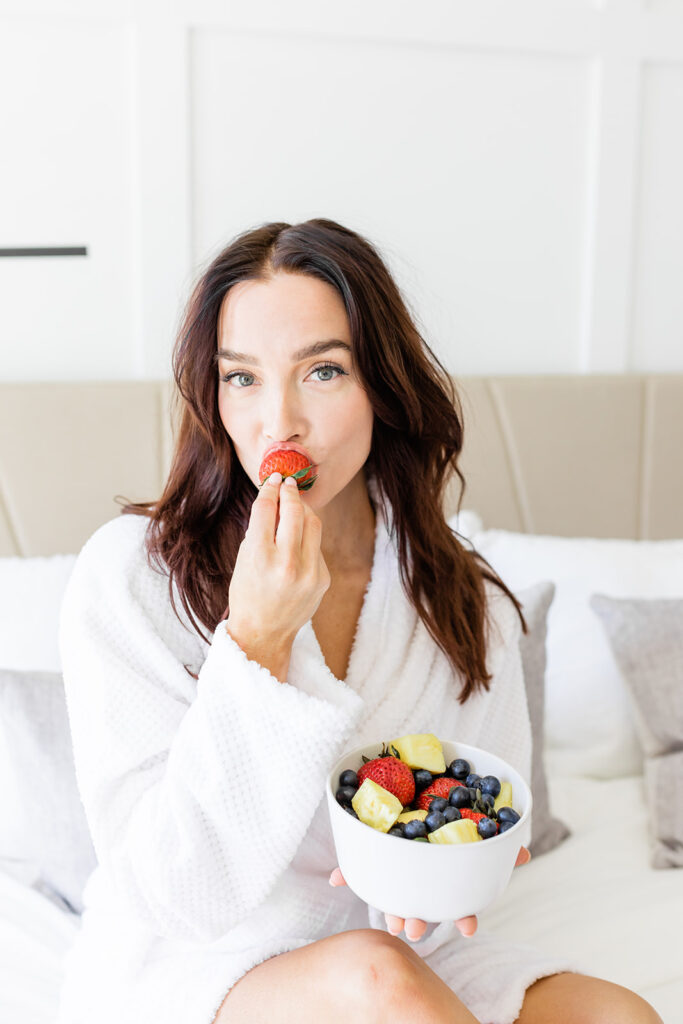 woman in robe holding bowl of fruit eating strawberry